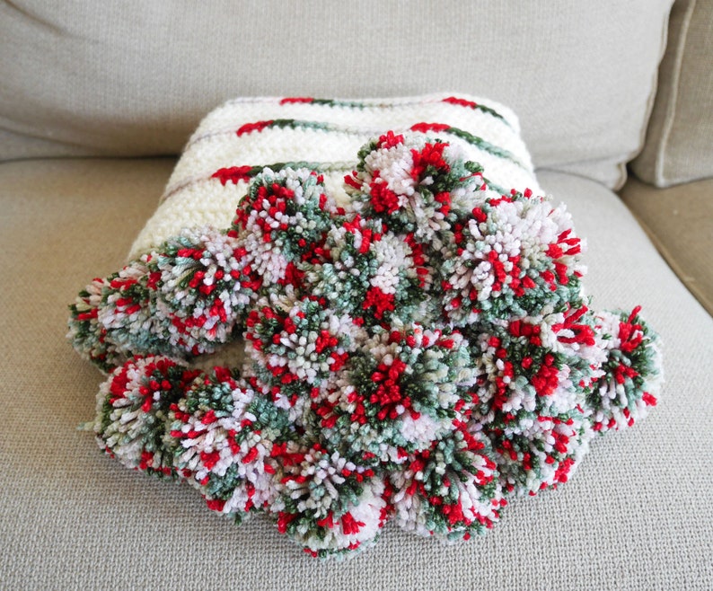 May include: A white crocheted throw blanket with green and red pom-poms. The blanket is made with a striped pattern and has a soft, fluffy texture.