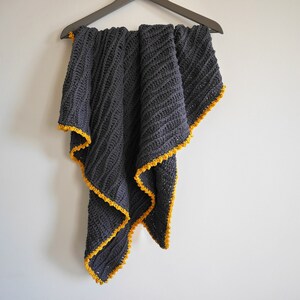 May include: A dark gray crocheted throw blanket with a yellow crocheted border. The blanket is hanging on a hanger against a white wall.