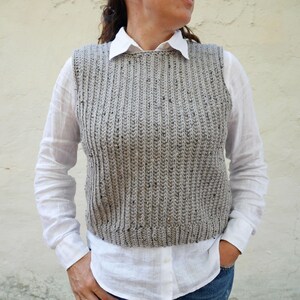 Peut inclure: Un gilet sans manches en tricot gris avec un motif moucheté. Le gilet a un col rond et une ourlet droit.