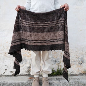 May include: A brown and beige crocheted shawl with a fringed edge. The shawl is being held up by a person wearing white trousers and brown shoes.