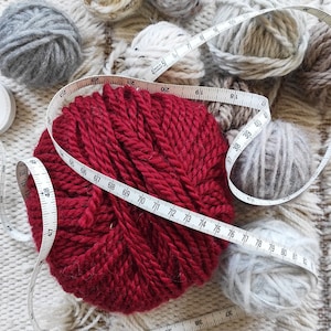 Puede incluir: Una bola de lana roja está envuelta con una cinta métrica blanca. La cinta indica de 45 a 82 pulgadas. Hay otras bolas de lana en el fondo, incluyendo grises, blancas y beige.