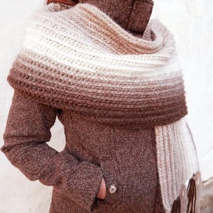 Könnte beinhalten: Ein brauner und weißer Ombre-Häkel-Schal mit Fransen. Der Schal wird um den Hals einer Person getragen, die eine braune Tweedjacke trägt.