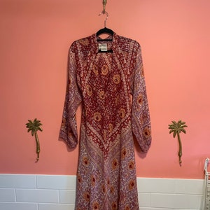 Puede incluir: Vestido de manga larga con un vibrante estampado floral en tonos rojos, rosas y dorados. El vestido está colgado de una percha negra contra una pared rosa. El vestido tiene un cuello alto y una falda fluida.