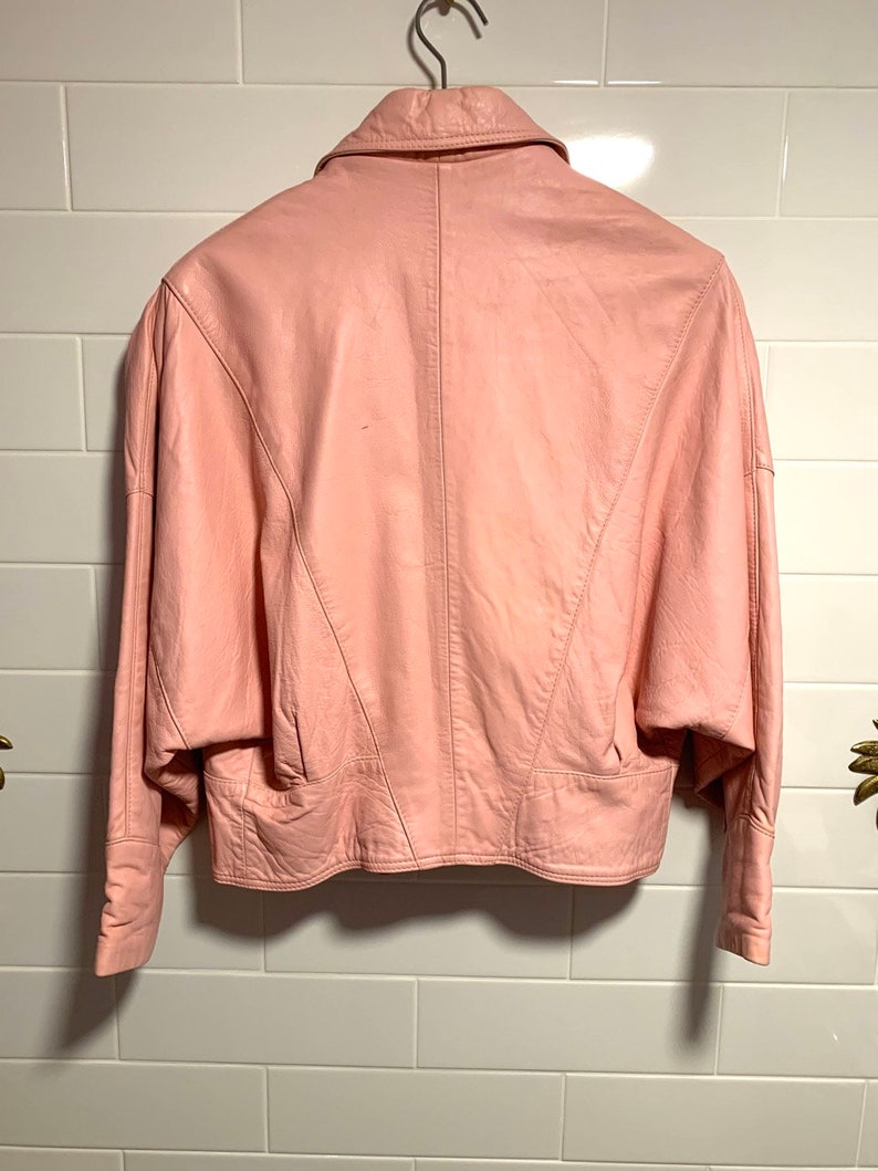 Puede incluir: Una chaqueta de cuero rosa con cuello y mangas largas. La chaqueta tiene una silueta ajustada y una longitud recortada.