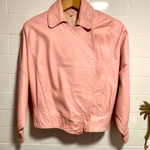 Puede incluir: Una chaqueta de cuero rosa con cuello y cierre de botones. La chaqueta tiene mangas largas y una longitud recortada.