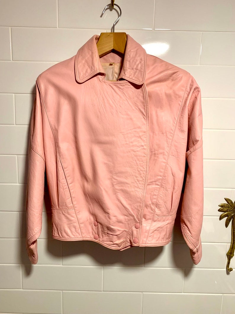 Puede incluir: Una chaqueta de cuero rosa con cuello y cierre de botones. La chaqueta tiene mangas largas y una longitud recortada.
