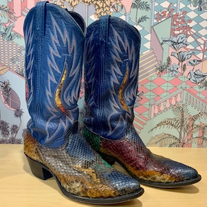 Könnte beinhalten: Ein Paar Cowboy-Stiefel. Die Schäfte sind aus blauem Leder mit goldenen Nähten. Der Fuß der Stiefel hat ein Schlangenmuster in Braun-, Grün- und Rottönen. Die Stiefel stehen auf einer Holzoberfläche.