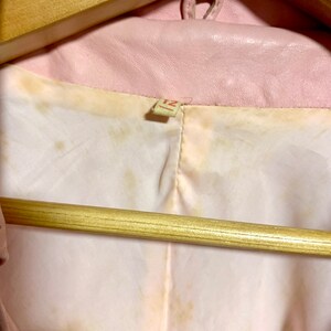 Puede incluir: Una chaqueta de cuero rosa colgada de una percha de madera marr&oacute;n. La chaqueta tiene una etiqueta que dice "12".