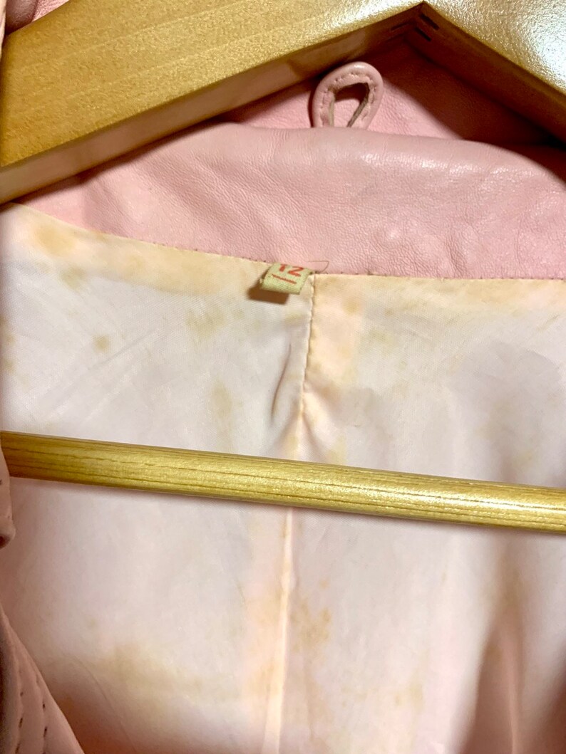 Puede incluir: Una chaqueta de cuero rosa colgada de una percha de madera marr&oacute;n. La chaqueta tiene una etiqueta que dice "12".