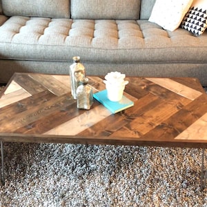 Puede incluir: Una mesa de centro de madera rústica con una tapa de patrón de chevron. La mesa tiene una mancha marrón oscura y está soportada por cuatro patas de horquilla. La mesa está decorada con tres botellas de vidrio plateadas y una maceta de cerámica blanca.