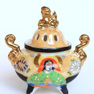 Può includere: Un bruciatore di incenso in ceramica con un coperchio dorato e tre gambe. Il bruciatore è decorato con un motivo floreale colorato e una figura di una donna in un abito verde e rosso.