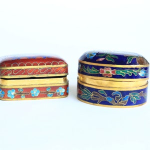 Op de afbeelding: Twee kleine, sierlijke, decoratieve doosjes met ingewikkelde bloemmotieven. Het doosje links is rood met blauwe en witte accenten. Het doosje rechts is blauw met gouden, groene en roze accenten.