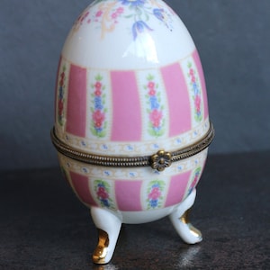 May include: A porcelain egg-shaped trinket box with a pink and white striped pattern and floral accents. The box has a hinged lid and sits on three gold-painted feet.