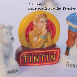 May include: Figurines of Tintin characters, including Tintin, Snowy, and a central figure in a yellow and red frame. The figurines are on white bases, with the text "Tintin" and "Milou" visible.