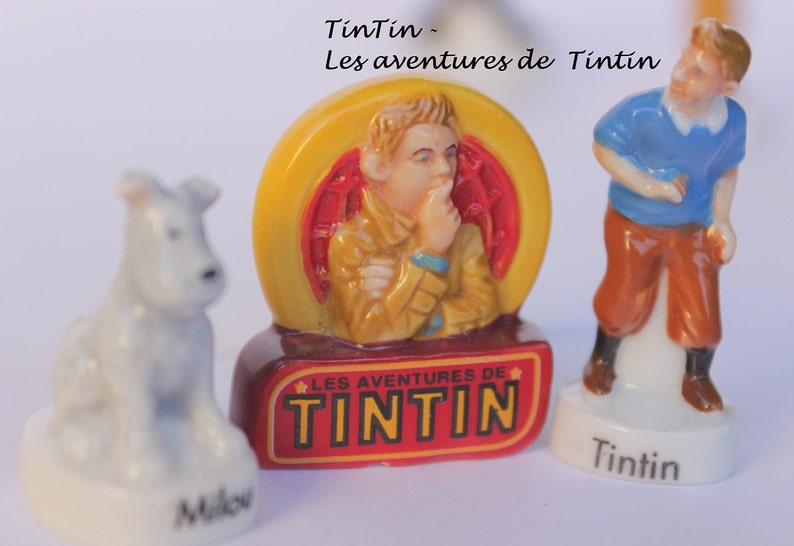 May include: Figurines of Tintin characters, including Tintin, Snowy, and a central figure in a yellow and red frame. The figurines are on white bases, with the text "Tintin" and "Milou" visible.
