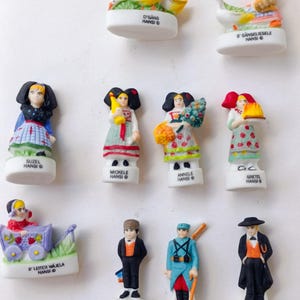 Puede incluir: Una colecci&oacute;n de peque&ntilde;as figuras de cer&aacute;mica blanca que representan personajes y escenas alsacianas. Las figuras presentan ropa y accesorios detallados, algunos sosteniendo objetos como flores o cestas. El texto en las bases incluye nombres y la palabra "HANSI".