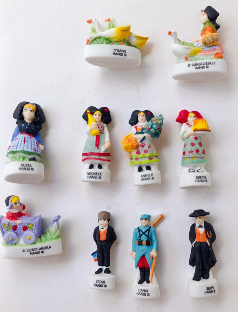 Puede incluir: Una colecci&oacute;n de peque&ntilde;as figuras de cer&aacute;mica blanca que representan personajes y escenas alsacianas. Las figuras presentan ropa y accesorios detallados, algunos sosteniendo objetos como flores o cestas. El texto en las bases incluye nombres y la palabra "HANSI".