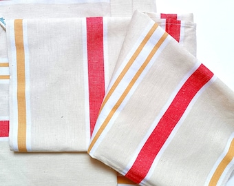Puede incluir: Conjunto de cinco toallas de cocina con rayas rojas y amarillas sobre un fondo beige. Las toallas están dobladas y dispuestas sobre una superficie blanca.