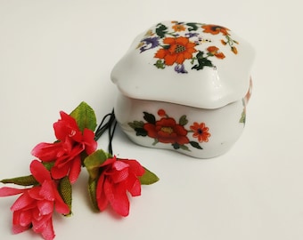 May include: A white porcelain trinket box with a floral design featuring orange, red, and purple flowers. The box has a lid and is decorated with a floral pattern.