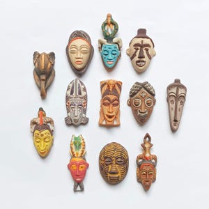 May include: A collection of twelve small, decorative wall masks featuring diverse facial expressions and intricate patterns. The masks are rendered in various colors including brown, yellow, red, blue, and white, showcasing distinct carved details.