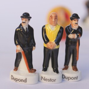May include: Three ceramic figurines of characters, each standing on a white base with a name. Two figures wear black suits and hats, one with a cane. The central figure wears a yellow vest. Names include "Dupont" and "Nestor".