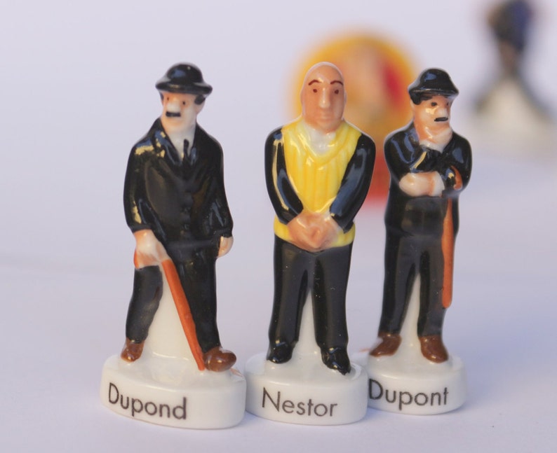 May include: Three ceramic figurines of characters, each standing on a white base with a name. Two figures wear black suits and hats, one with a cane. The central figure wears a yellow vest. Names include "Dupont" and "Nestor".