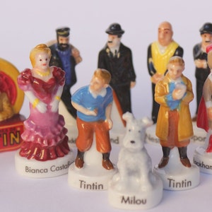 May include: A collection of ceramic figurines featuring characters from the Tintin comic series. The figurines depict Tintin, Snowy, Captain Haddock, and other characters. The figurines are painted in bright colors and stand on white bases.
