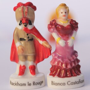 May include: Two porcelain figurines. One is a figure in a tan outfit with a red cape and hat, labeled "Rackham le Rouge." The other is a woman in a pink and red dress, labeled "Bianca Castafiore." Both figures are on white bases.