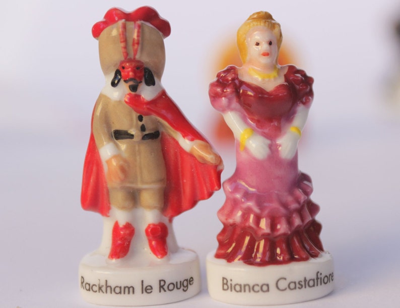 May include: Two porcelain figurines. One is a figure in a tan outfit with a red cape and hat, labeled "Rackham le Rouge." The other is a woman in a pink and red dress, labeled "Bianca Castafiore." Both figures are on white bases.
