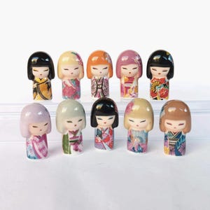 Könnte beinhalten: Eine Sammlung von elf japanischen Kokeshi-Puppen. Jede Puppe hat eine einzigartige Frisur und ein Kimono-Design in verschiedenen Farben, darunter Schwarz, Rosa, Orange und Gold. Die Puppen sind in zwei Reihen angeordnet und zeigen ihre detaillierte Handwerkskunst.