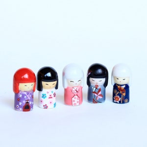 Könnte beinhalten: Fünf kleine, dekorative japanische Kokeshi-Puppen. Jede Puppe hat einen abgerundeten Kopf mit einem bemalten Gesicht und einen zylindrischen Körper. Die Puppen sind in verschiedenen Farben bemalt, darunter Rot, Schwarz, Weiß, Rosa und Blau, mit floralen und geometrischen Mustern.