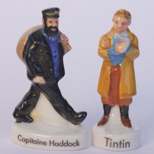 May include: Two ceramic figurines of characters from The Adventures of Tintin. One figure is Captain Haddock, wearing a black coat and carrying a bag. The other is Tintin, in a tan coat. Each figure is on a white base with the character's name.
