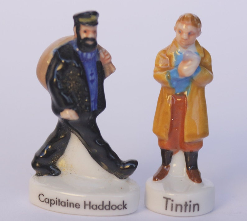 May include: Two ceramic figurines of characters from The Adventures of Tintin. One figure is Captain Haddock, wearing a black coat and carrying a bag. The other is Tintin, in a tan coat. Each figure is on a white base with the character's name.