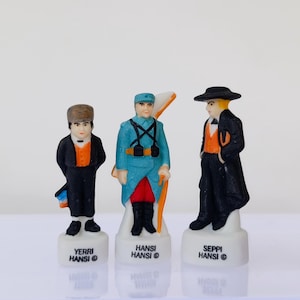 Puede incluir: Tres peque&ntilde;as figuras de cer&aacute;mica de hombres con trajes tradicionales. Las figuras est&aacute;n sobre bases blancas con el texto "YERRI HANSI", "HANSI HANSI" y "SEPPI HANSI". Las figuras son de color negro, azul, naranja y blanco.