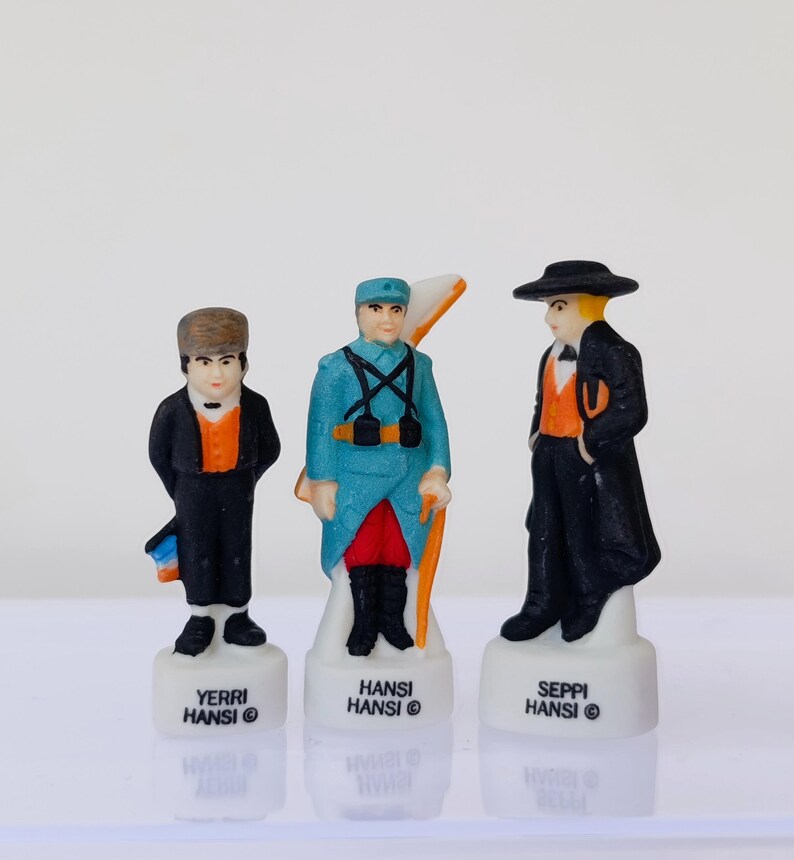 Puede incluir: Tres peque&ntilde;as figuras de cer&aacute;mica de hombres con trajes tradicionales. Las figuras est&aacute;n sobre bases blancas con el texto "YERRI HANSI", "HANSI HANSI" y "SEPPI HANSI". Las figuras son de color negro, azul, naranja y blanco.