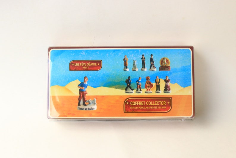 May include: A collector's box featuring miniature porcelain figurines. The box has a blue background with various characters and the text "COFFRET COLLECTOR". The figurines include Tintin and Snowy, and other characters.