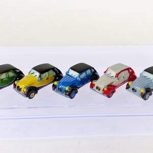 以下が含まれることがあります： さまざまな色の5台のミニチュアおもちゃの車が並んでいます。車は黒い屋根と、緑、黄色、青、赤と白、水色のボディカラーをしています。透明な長方形の表面に車が置かれています。