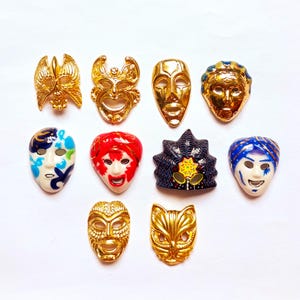 May include: A collection of ten small decorative mask pins or brooches arranged in three rows. The set includes various designs, featuring metallic gold finishes, as well as painted masks in blue, white, red, and black with intricate facial details.
