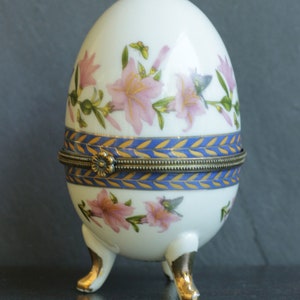 May include: A white porcelain egg-shaped trinket box with a gold and blue trim and floral design. The box has three gold-painted feet.