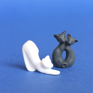 以下が含まれることがあります： 白いセラミック製の猫の置物と黒いセラミック製の猫の置物の2つ。白い猫は遊び心のあるポーズで丸まっており、黒い猫はよりリラックスしたポーズをとっています。両方の置物は青い背景の上にあります。