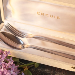 May include: A white box with the word "ERCUIS" printed on the lid. Inside the box are two silver pieces of cutlery: a fork and a spoon. The box is lined with white satin.
