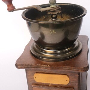 May include: A vintage wooden coffee grinder with a metal crank and a drawer for collecting coffee grounds. The grinder has a dark brown wooden base and a metal top with a black interior.