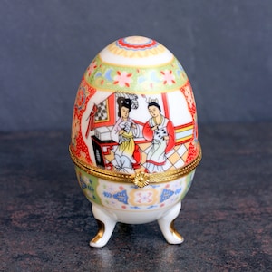 May include: A porcelain egg-shaped trinket box with a gold trim and three gold feet. The box is decorated with a colorful floral pattern and a scene of two women in traditional Chinese clothing.
