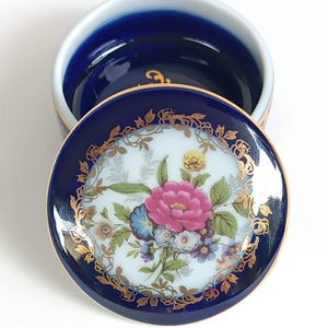 May include: A small, round porcelain trinket box with a blue and gold floral design. The lid features a white background with a pink and yellow floral pattern surrounded by a gold border. The box is cobalt blue with a gold trim.