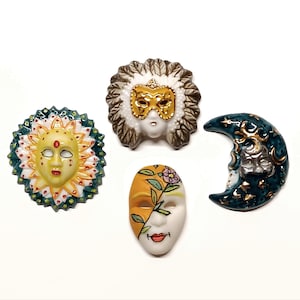 Pode incluir: Quatro máscaras decorativas de cerâmica: um sol com rosto amarelo e detalhes vermelhos, uma máscara com máscara dourada e cocar de penas, uma meia máscara com seções laranja e branca e uma lua crescente com rosto.