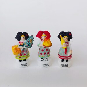 Puede incluir: Tres peque&ntilde;as figuras decorativas de mujeres con trajes tradicionales. Cada figura tiene un peinado &uacute;nico y sostiene diferentes objetos, incluyendo un ramo, un fardo y una botella. Las figuras est&aacute;n sobre bases blancas con los nombres "Annele Hansi", "Gretel Hansi" y "Mickele Hansi".