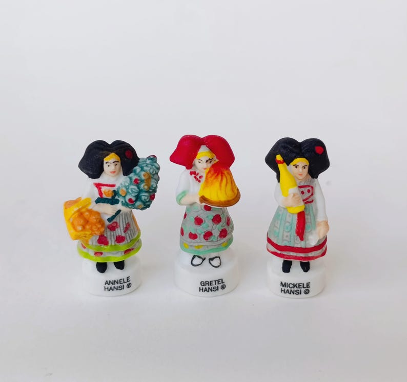 Puede incluir: Tres peque&ntilde;as figuras decorativas de mujeres con trajes tradicionales. Cada figura tiene un peinado &uacute;nico y sostiene diferentes objetos, incluyendo un ramo, un fardo y una botella. Las figuras est&aacute;n sobre bases blancas con los nombres "Annele Hansi", "Gretel Hansi" y "Mickele Hansi".