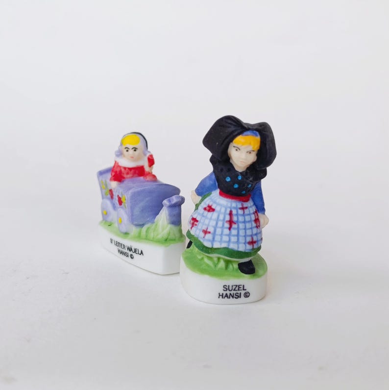 Puede incluir: Dos peque&ntilde;as figuras de cer&aacute;mica. Una representa a una persona con abrigo rojo montada en un carro azul con detalles florales. La otra es una persona con un gorro negro tradicional y un vestido azul. Las figuras est&aacute;n sobre bases blancas con texto.