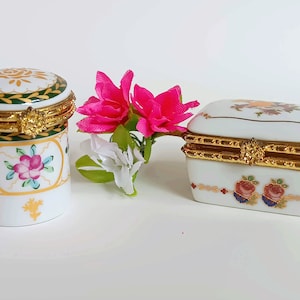 May include: Two porcelain trinket boxes with floral designs. One is round and the other is rectangular. Both have gold trim and hinges.