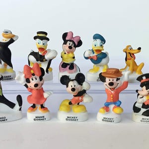 以下が含まれることがあります： ミッキーマウス、ミニーマウス、ドナルドダック、グーフィーなど、10体のディズニーキャラクターのフィギュアのコレクション。鮮やかな色のフィギュアは、キャラクター名が書かれた白い台座の上に立っています。